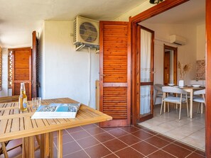 A room with a wooden table, a bottle, and a glass, a wooden door, a white wall, and an air conditioning unit. - Fortuna by Interhome (Villasimius)
