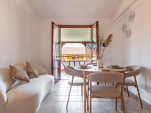 A modern living room with a sofa, dining table, chairs, and a view of a traditional building through an open door. - Fortuna by Interhome (Villasimius)