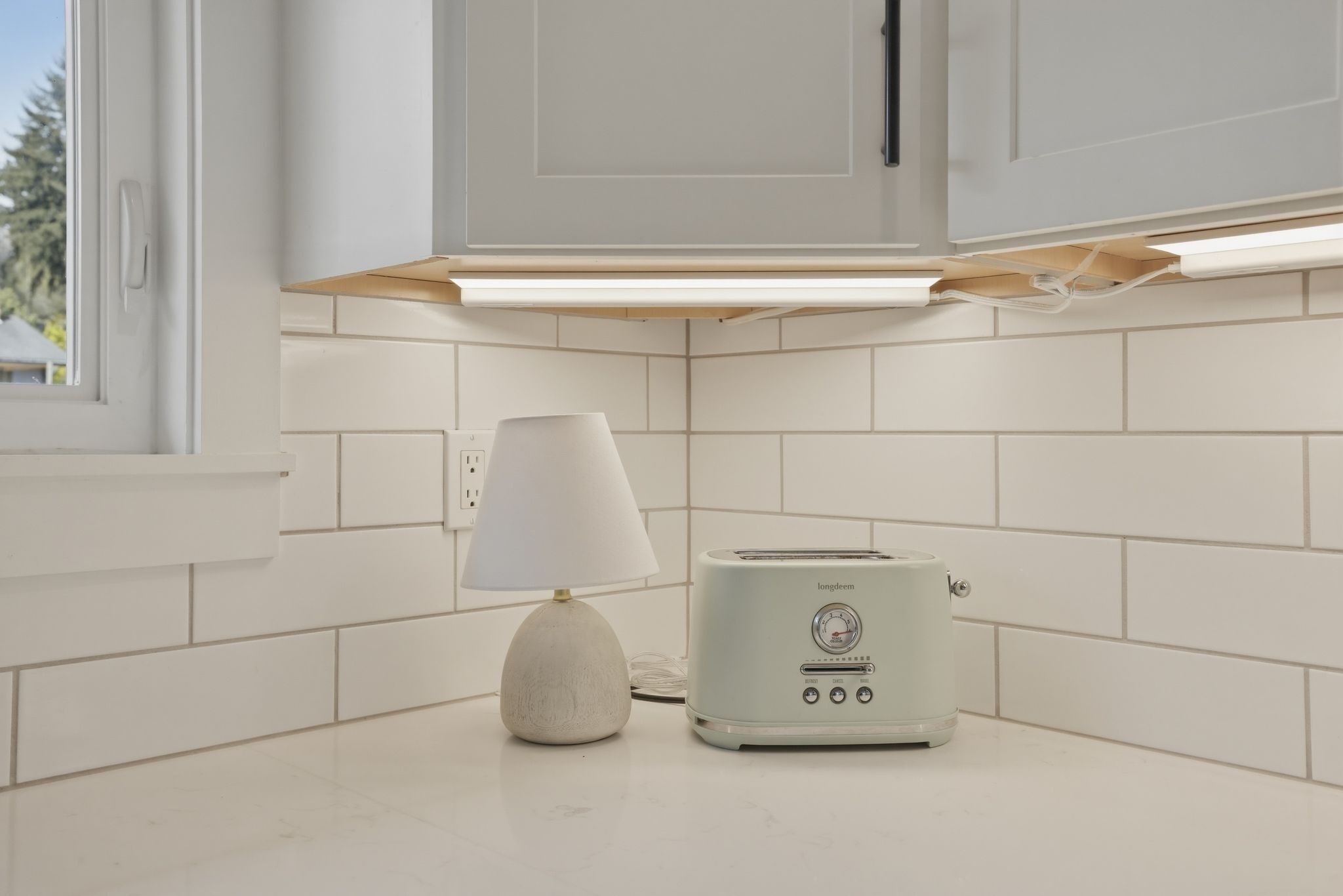Une cuisine avec des meubles blancs, un plan de travail blanc, une lampe blanche et un grille-pain blanc.