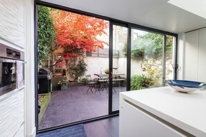 A modern kitchen with a sliding glass door leading to a patio with a red-leaved tree and outdoor furniture. - Elegant Family Home, Kensington (London)