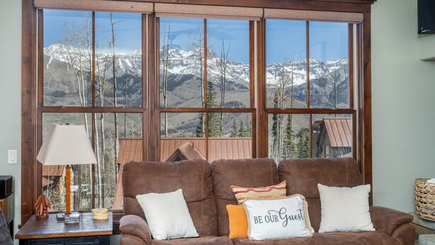 A brown sofa with three pillows, a lamp, and a view of snow-capped mountains through large windows. - Slopeside Reserve 2 Bedroom Condo (Telluride)