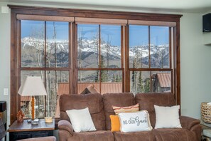 A brown sofa with three pillows, a lamp, and a view of snow-capped mountains through large windows. - Slopeside Reserve 2 Bedroom Condo (Telluride)