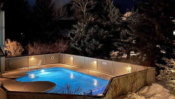 A heated outdoor swimming pool surrounded by snow, with a wooden deck and a chair, set against a mountainous backdrop.