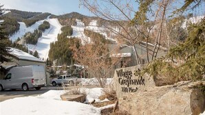 A sign for Village Greyhawk with a view of a snowy mountain and a parking area.