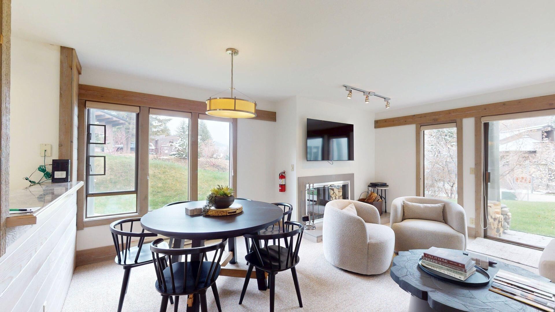 A spacious living area with a round table, chairs, a fireplace, a flat-screen TV, and large windows.