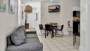 Ein modernes Wohnzimmer mit grauem Sofa, einem Essbereich mit Tisch und Stühlen, einem an der Wand befestigten Fernseher und einer Küchenzeile mit Geräten.
