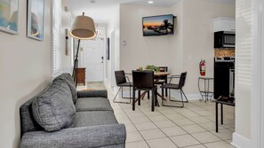 A modern living room with a grey sofa, a dining area with a table and chairs, a TV mounted on the wall, and a kitchen area with appliances.