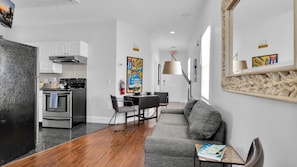 A modern kitchen with stainless steel appliances, a dining area with a black table and chairs, and a gray sofa in a bright living space.