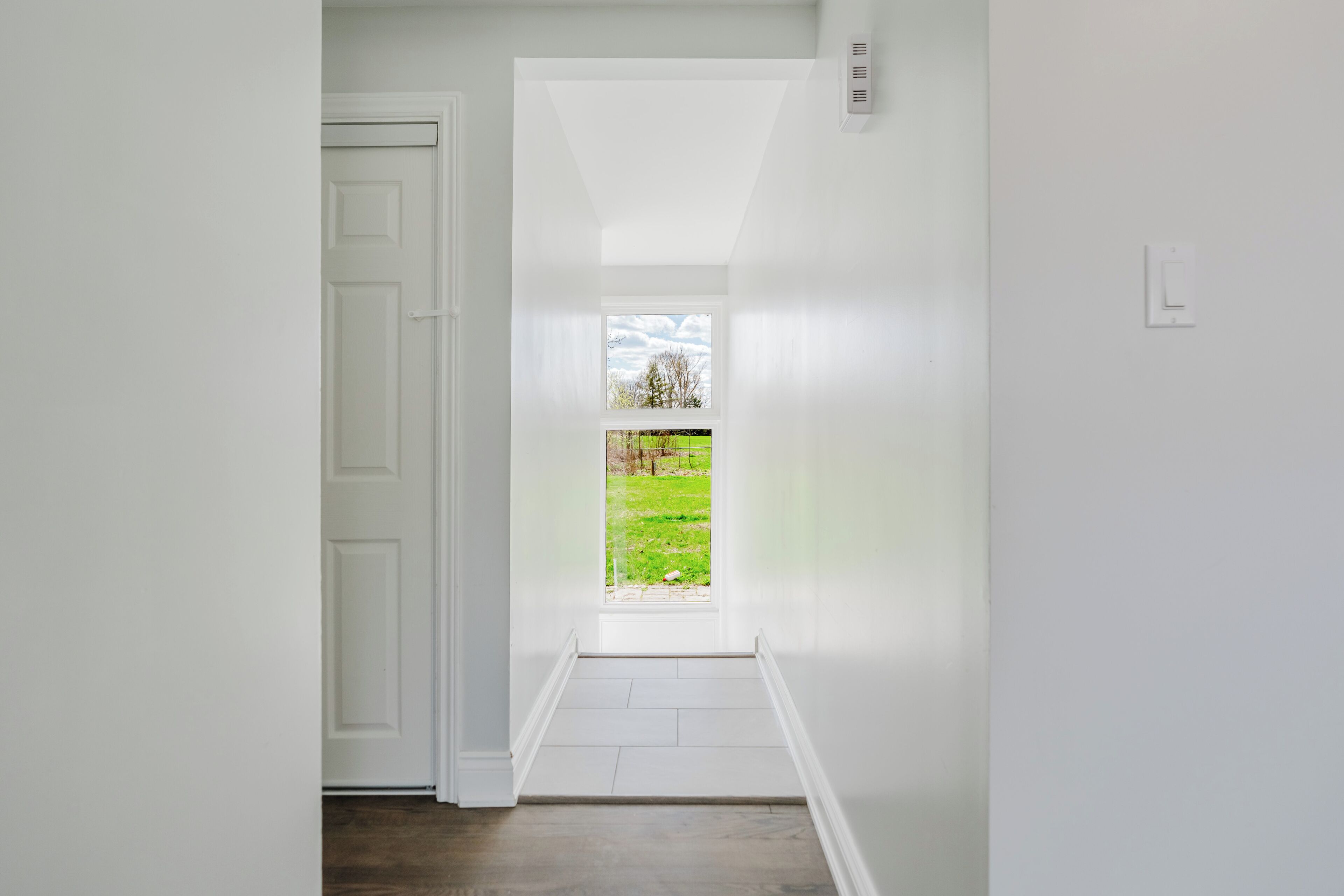 Un pasillo con una puerta que da a una habitación, una ventana con vista a un campo verde y una pared blanca.