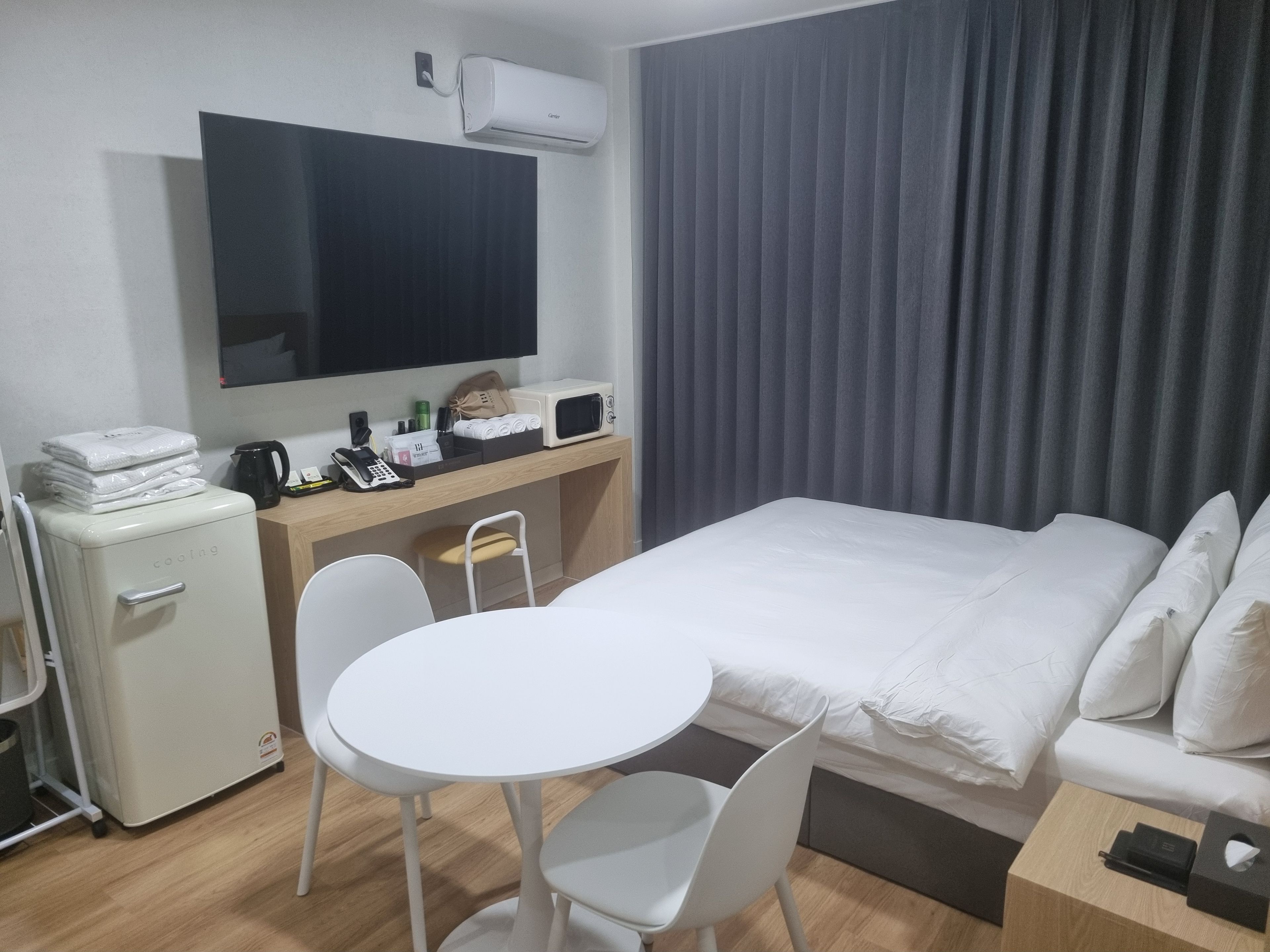 Ein Hotelzimmer mit einem Bett, einem Schreibtisch mit Computer und Telefon, einer kleinen Kühlschrank, einem Fernseher und einem Fenster mit Vorhängen.