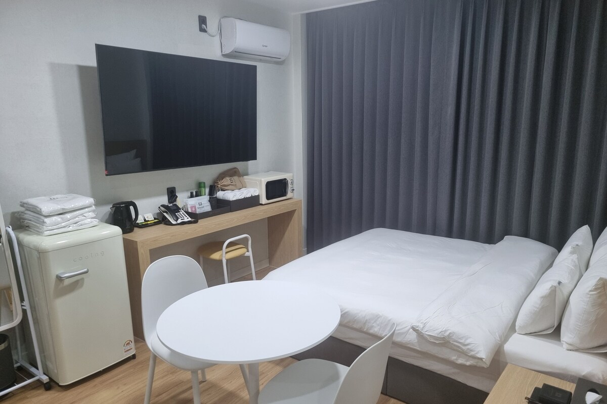 A hotel room with a bed, a desk with a computer and a phone, a small fridge, a TV, and a window with curtains.