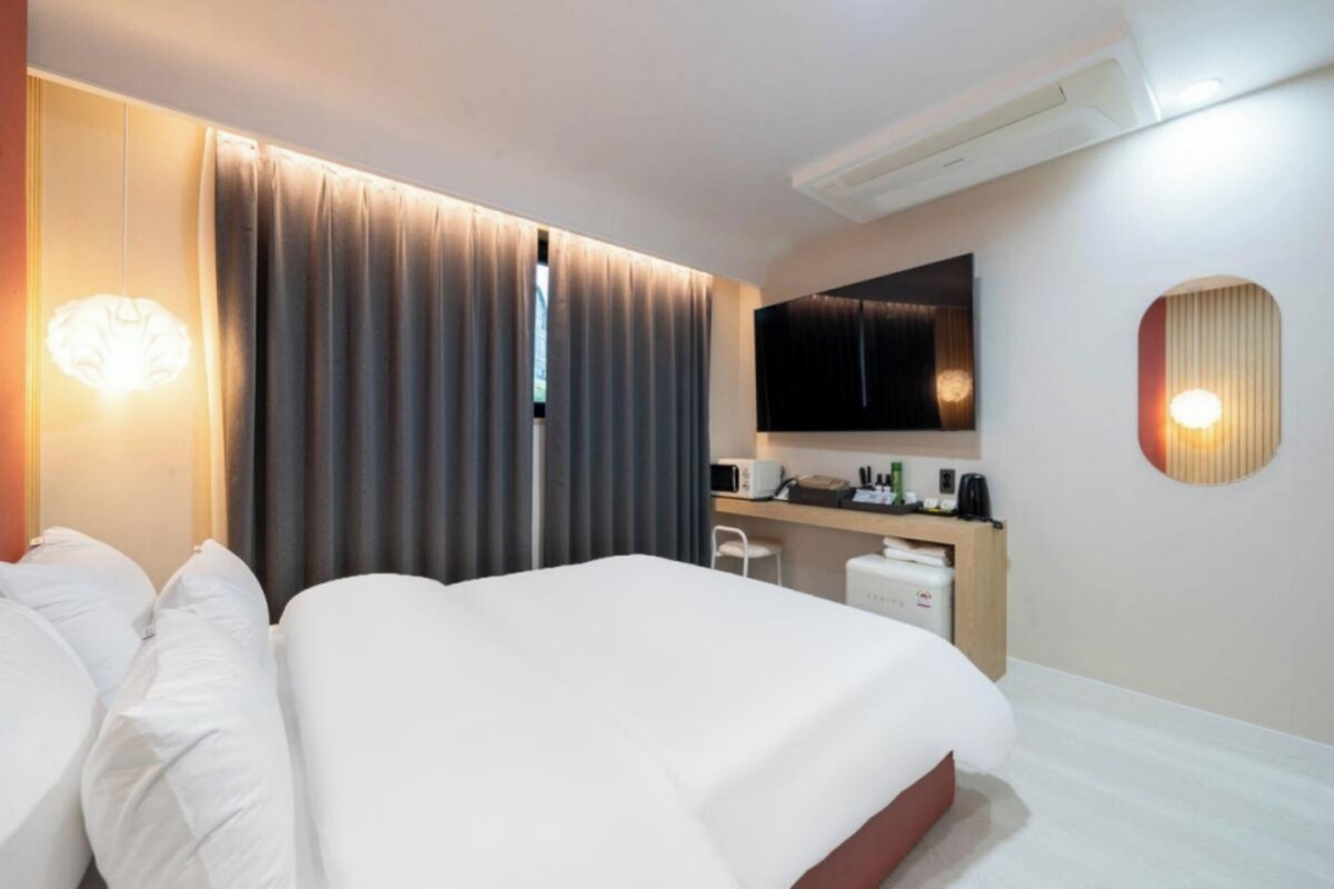 A hotel room with a bed, a desk, a flat-screen TV, and a wall-mounted light fixture.
