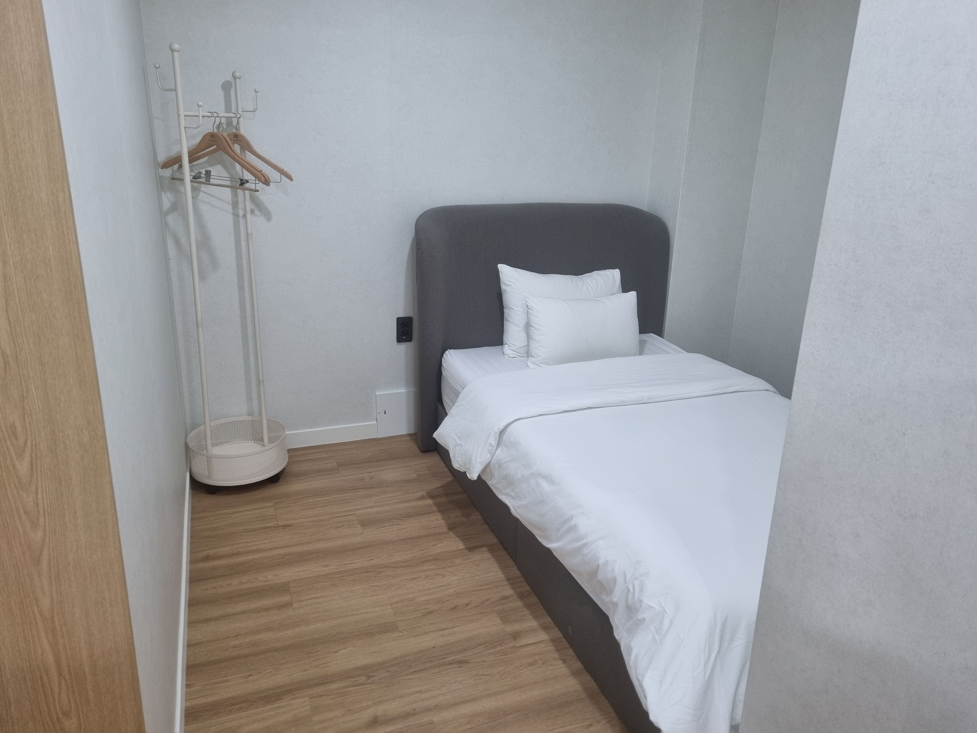 A small, neatly arranged bedroom with a single bed, a bedside table, a hanging clothes rack, and a floor lamp.