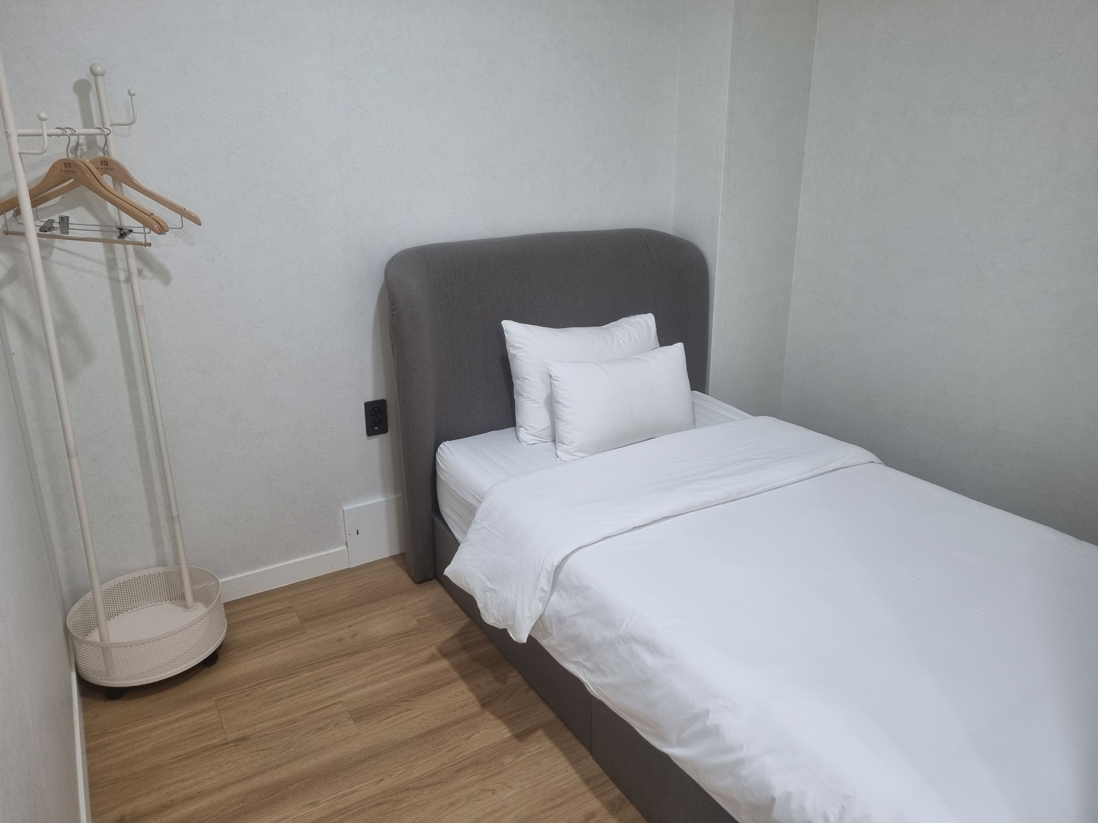 A single bed with a grey headboard and white bedding in a room with wooden flooring and a white wall.