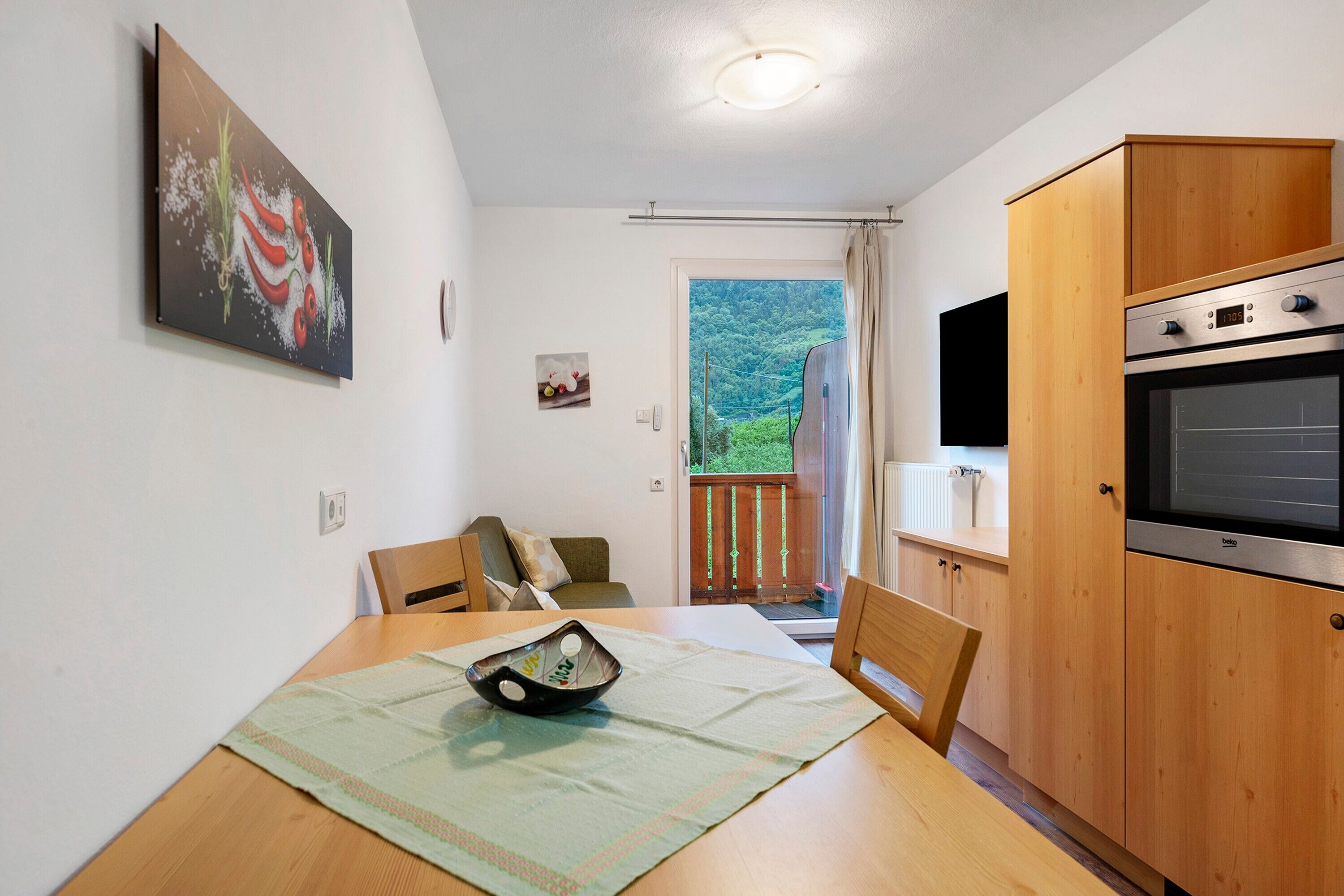 A kitchen with wooden cabinets, a dining table with chairs, a painting on the wall, and a view through the door.