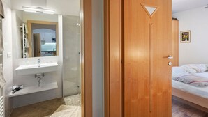 Een slaapkamer met een houten deur, een bed met gestreept beddengoed, een witte muur met een ingelijst schilderij, en een badkamer met een witte wastafel en spiegel.