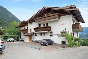 Ein zweistöckiges Gebäude mit Balkon, ein Auto davor und ein Berg im Hintergrund.