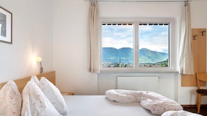 Un dormitorio con cama, almohadas, una ventana con vista a la montaña, un radiador y una silla.