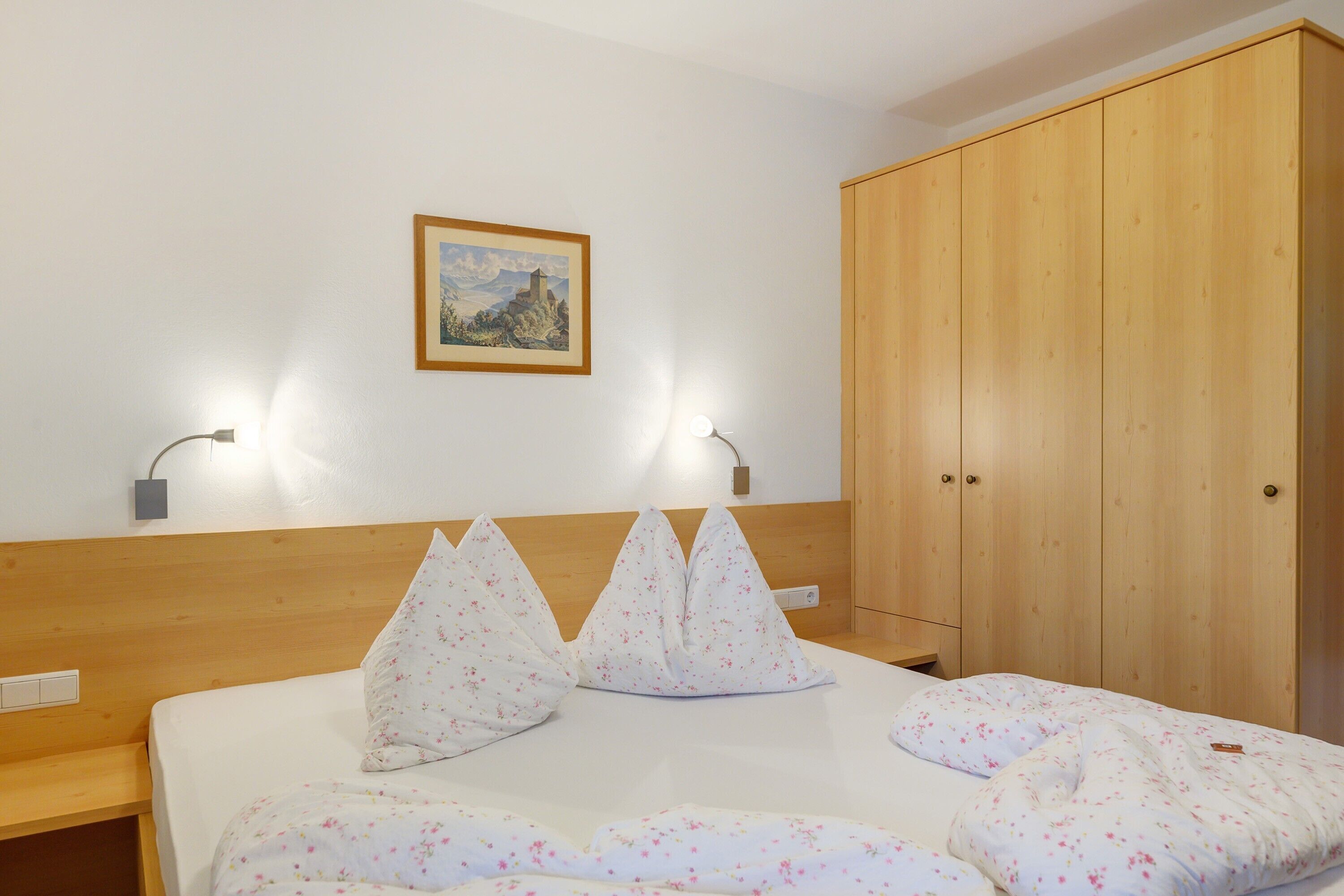 Une chambre d’hôtel avec une tête de lit en bois, un lit recouvert d’une literie à motifs floraux, une armoire en bois et un tableau encadré accroché au mur.
