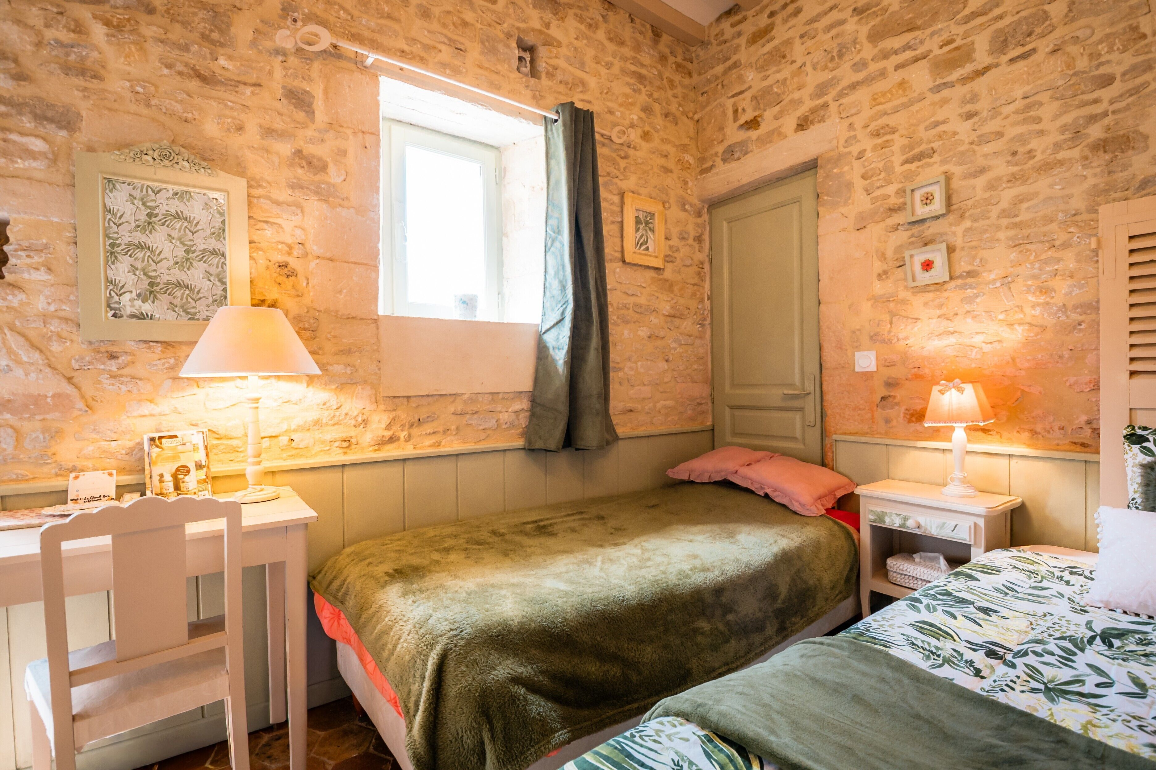 Une pièce aux murs de pierre, un lit recouvert d’une couverture verte, un bureau avec une lampe et une fenêtre avec des rideaux.