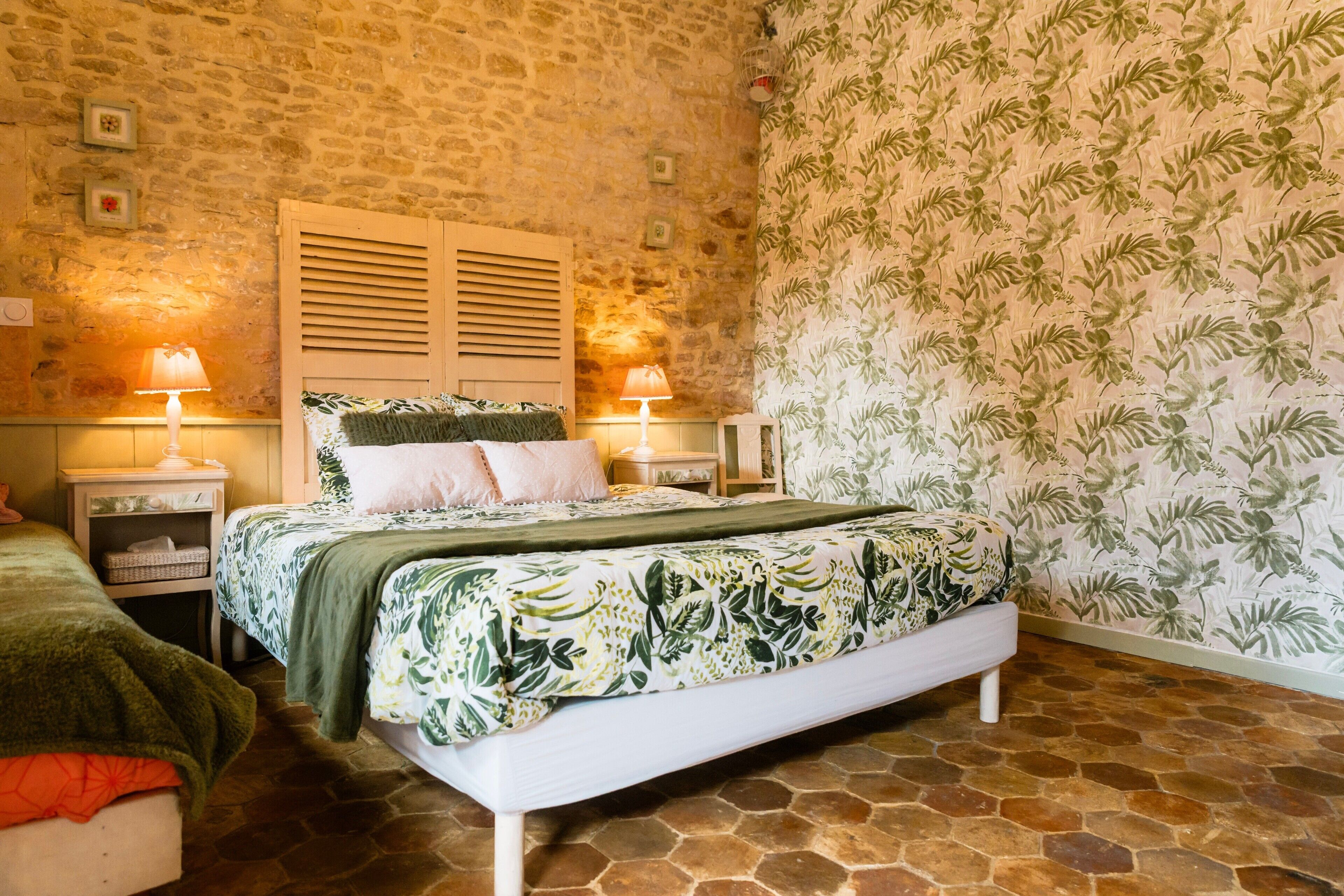 Quarto com parede de pedra, cama com colcha florida, duas luminárias de cabeceira e papel de parede estampado.