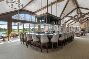 A spacious bar area with a large TV screen, multiple seating arrangements, and large windows offering views of the outdoors. - 27523 Medallion Ave., The Peninsula ~ (Millsboro)