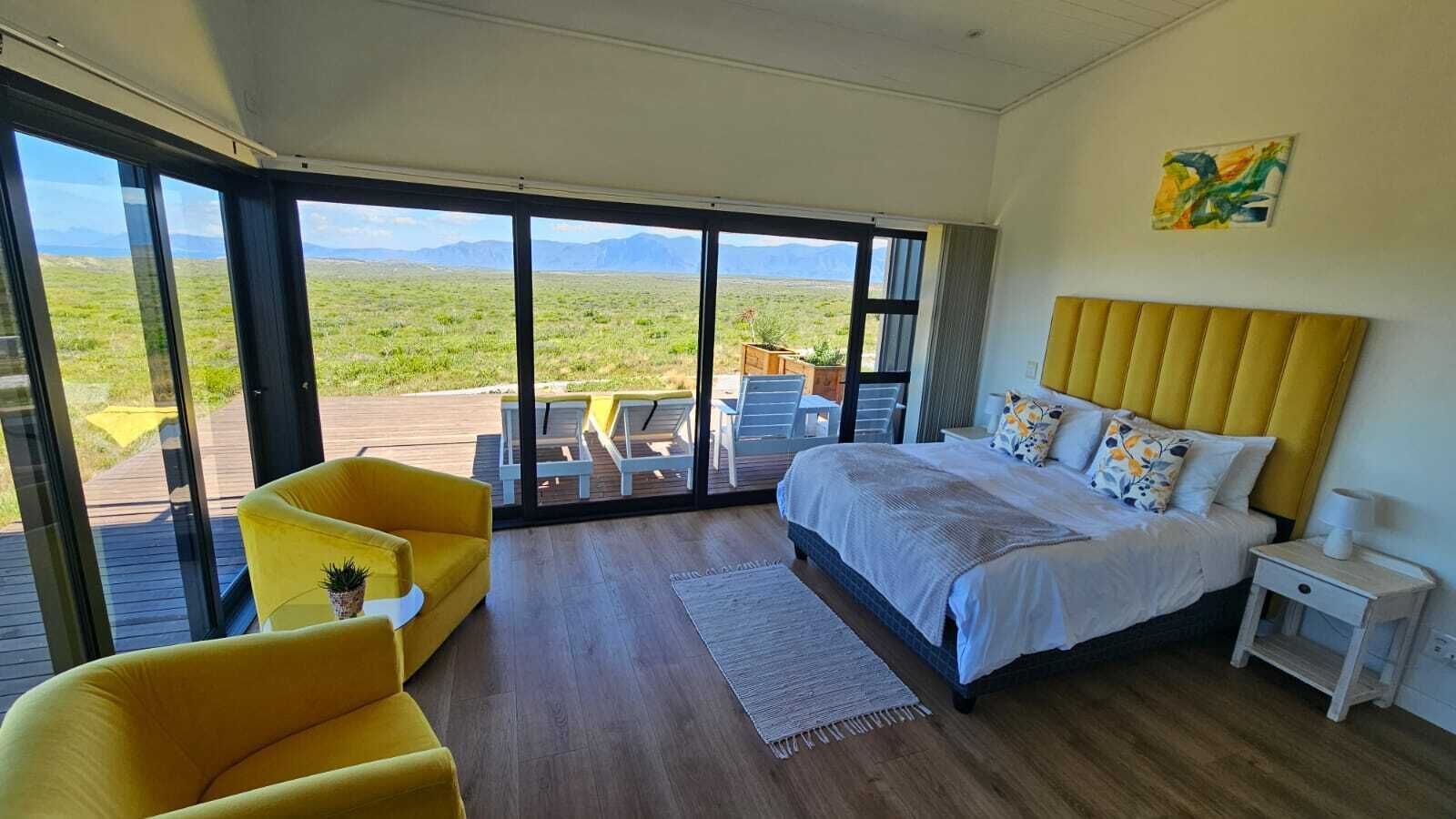 A bedroom with a bed, a yellow armchair, a nightstand, and a view of a grassy landscape through large windows.