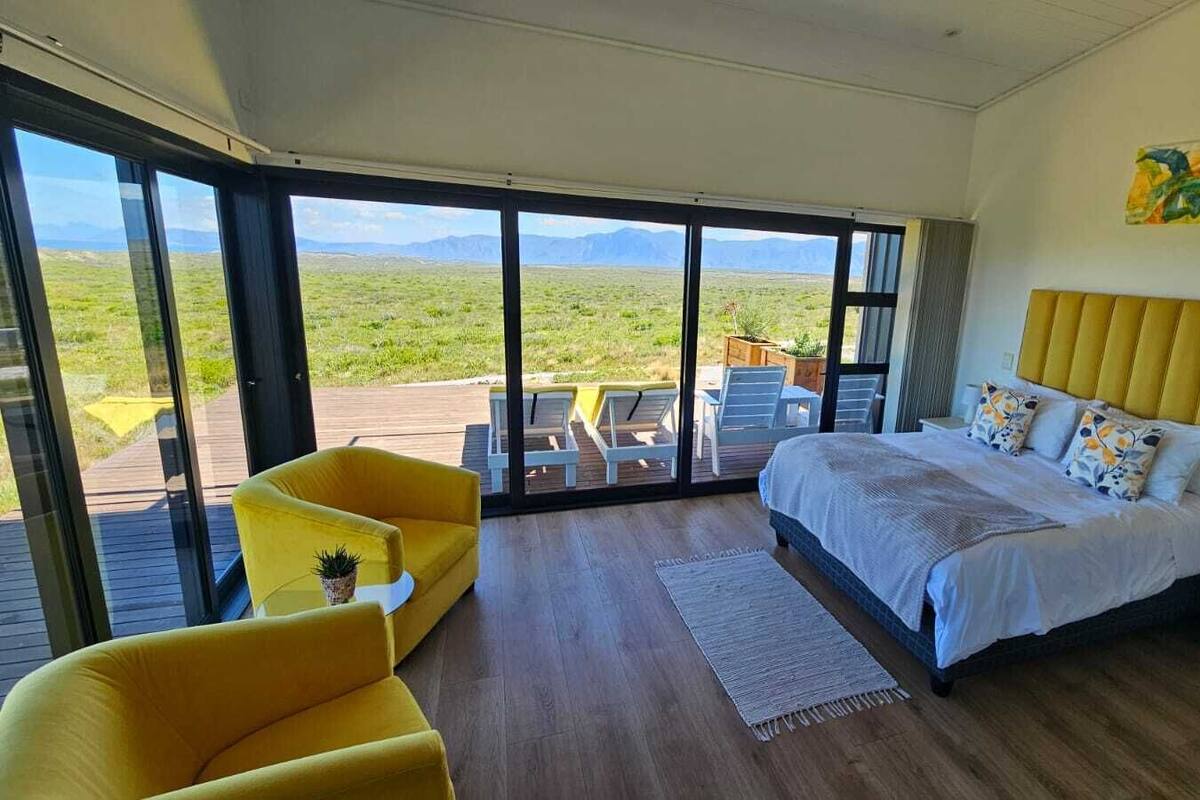 A bedroom with a bed, a yellow armchair, a nightstand, and a view of a grassy landscape through large windows.