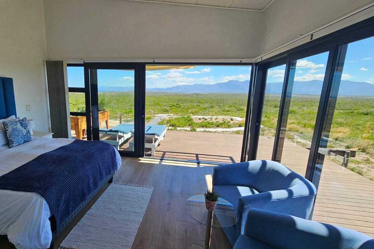 A bedroom with a large bed, a view of the outdoors, and a balcony with seating.