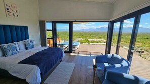 A bedroom with a large bed, a view of the outdoors, and a balcony with seating. - Walker Bay Dunes Lodge (Stanford)
