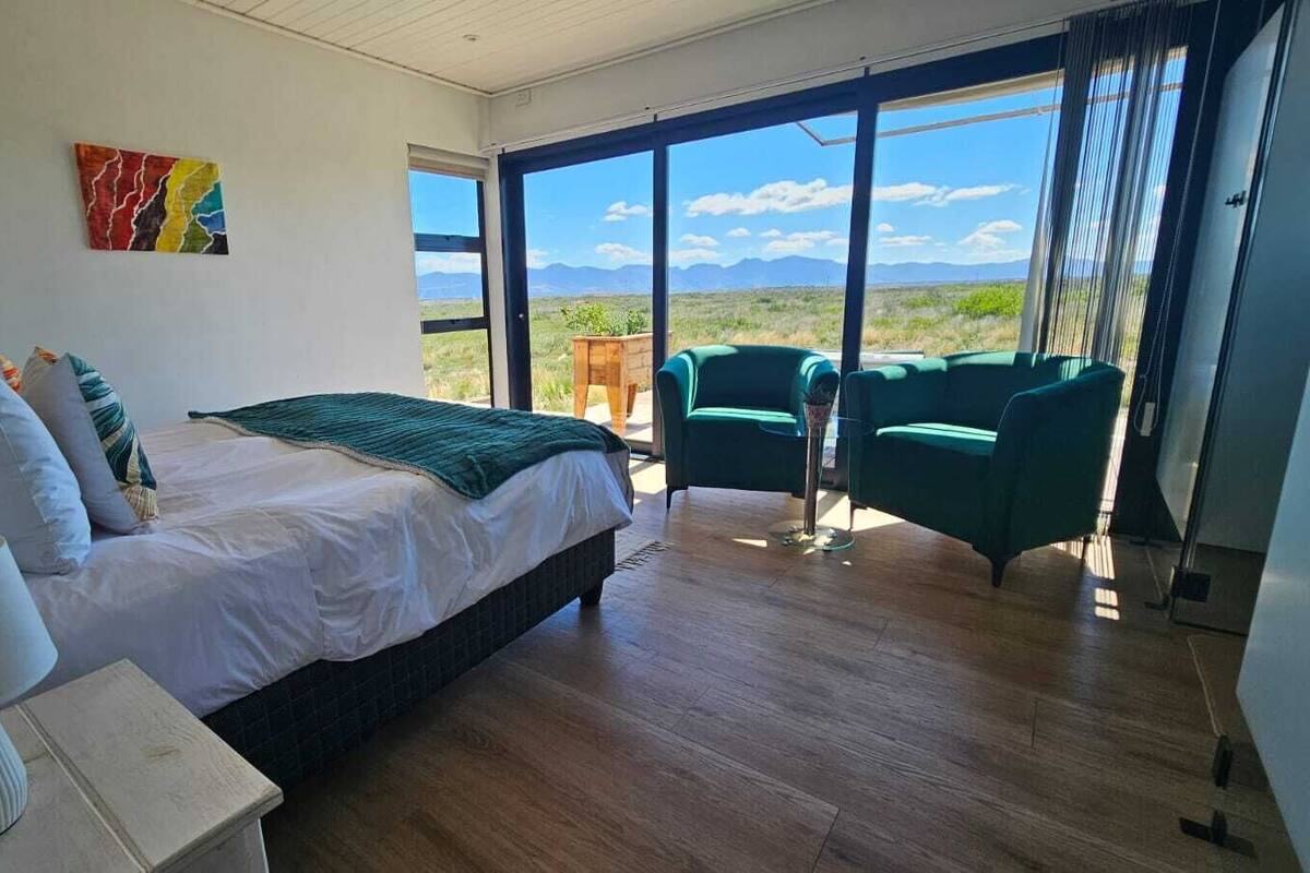 A bedroom with a bed, bedside table, lamp, and a view of the outdoors through large windows.