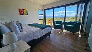 A bedroom with a bed, bedside table, lamp, and a view of the outdoors through large windows. - Walker Bay Dunes Lodge (Stanford)
