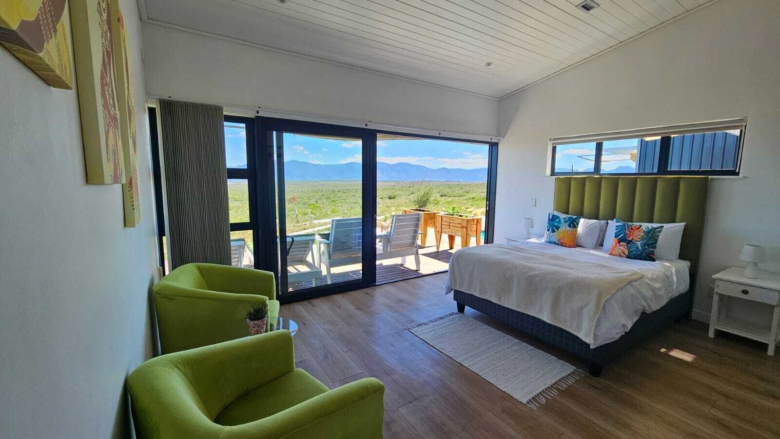 A bedroom with a bed, a green armchair, a nightstand, and a view of a grassy landscape through a sliding door.