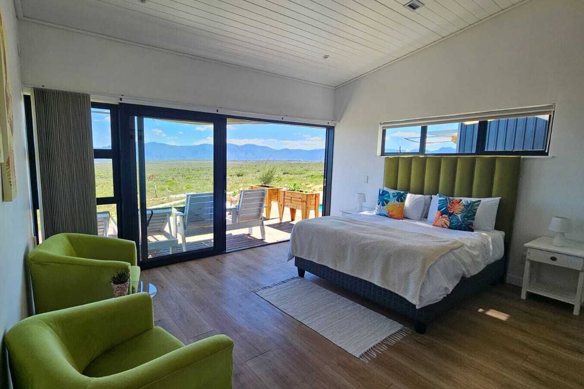 A bedroom with a bed, a green armchair, a nightstand, and a view of a grassy landscape through a sliding door.