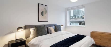 A bedroom with a bed, bedside table, lamp, dresser, window, and a framed picture on the wall.