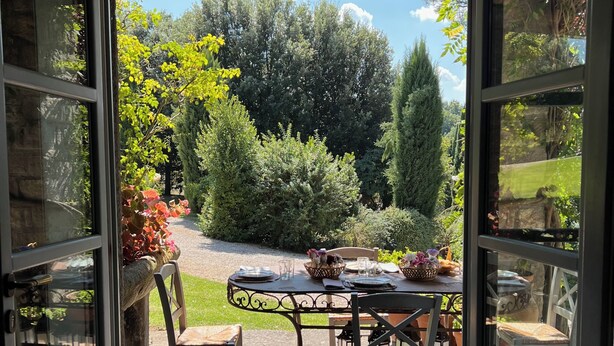 An open patio with a table set for dining, surrounded by chairs, and a view of a garden with trees and a lawn. - Villa Rossi Danielli (Lazio)