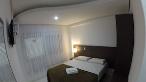 A hotel room with a bed, a nightstand, a lamp, a TV, and a window with blinds.