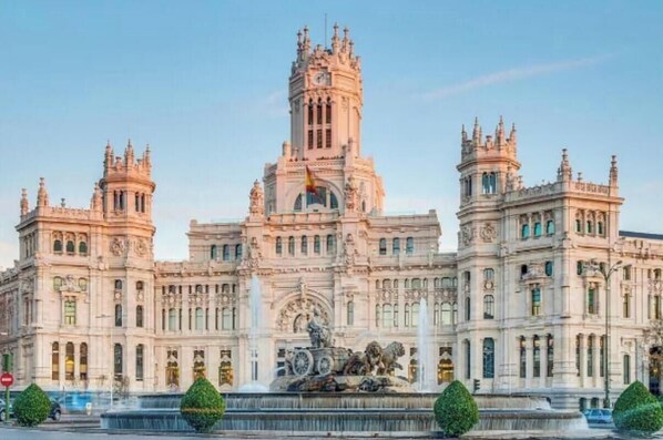 Un grande e decorato edificio con una torre centrale e due torri più piccole, circondato da una fontana e siepi curate.