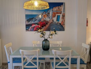 A dining area with a glass table, white chairs, a vase with flowers, and a painting of a coastal scene.