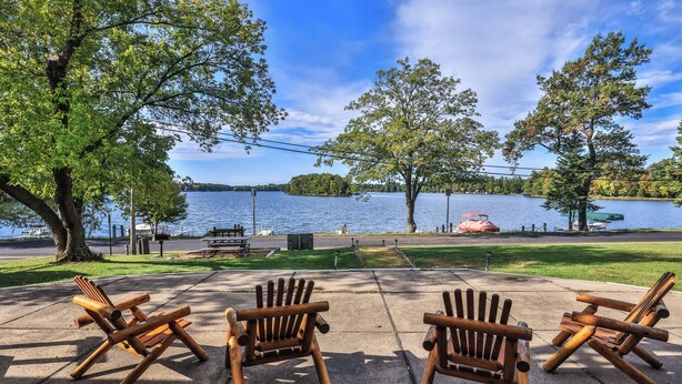 A lakeside view with three wooden Adirondack chairs facing the water, a picnic table, and a grassy area with trees.
