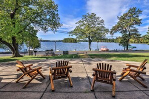 Vista del lago con tres sillas de madera tipo Adirondack orientadas al agua, una mesa de picnic y un área verde con árboles.