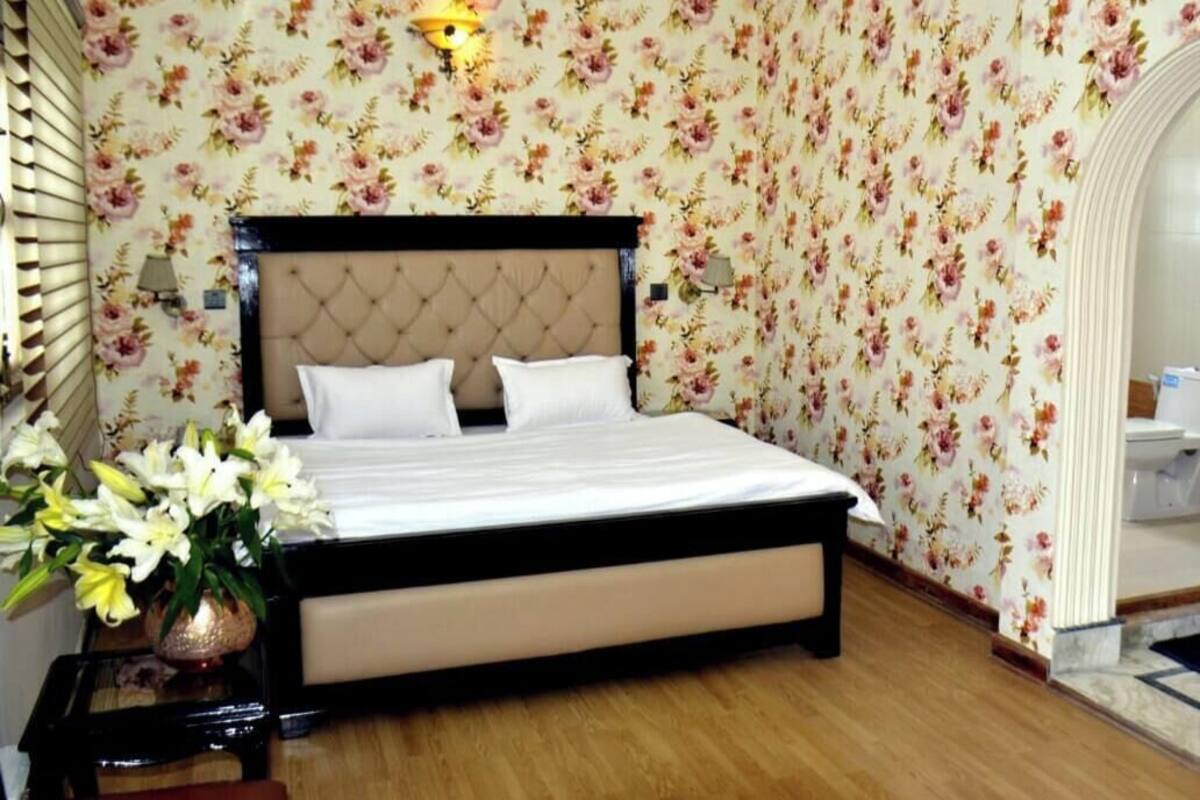 A bedroom with a bed, floral wallpaper, a nightstand with a vase of flowers, and a wooden floor.