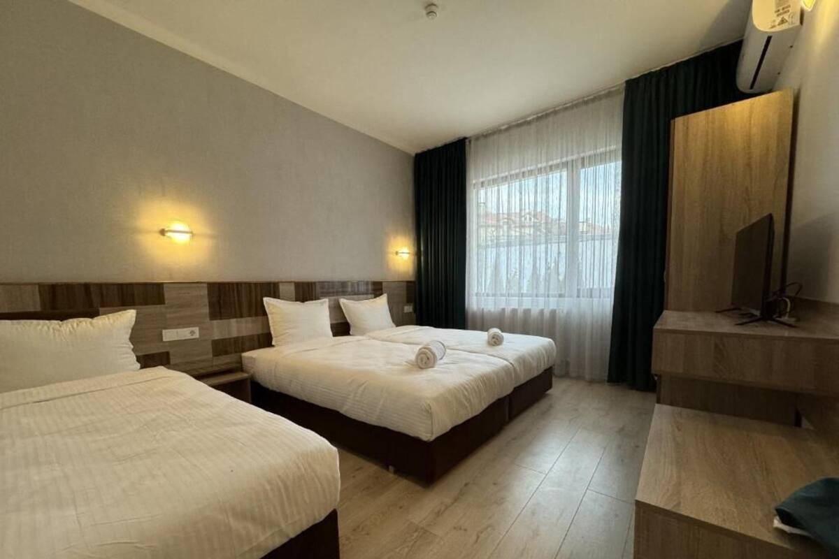 A hotel room with two beds, a wooden headboard, a large window with curtains, a wooden desk, and a mirror.