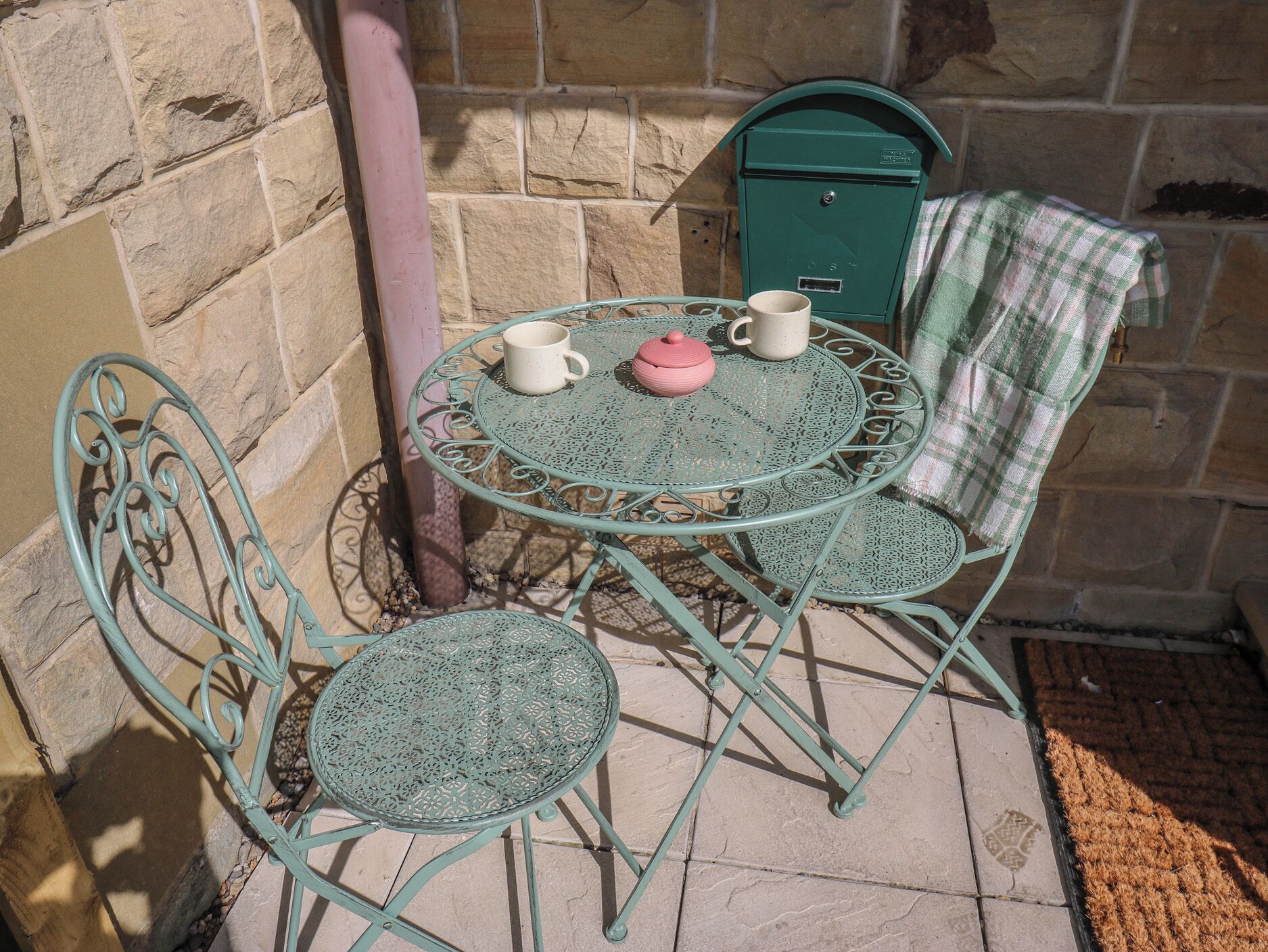 A metal outdoor table with two chairs, a green mailbox, and a brick wall.