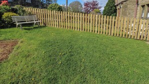A grassy backyard with a wooden fence, a bench, and a stone building. - Underview (Holmfirth)