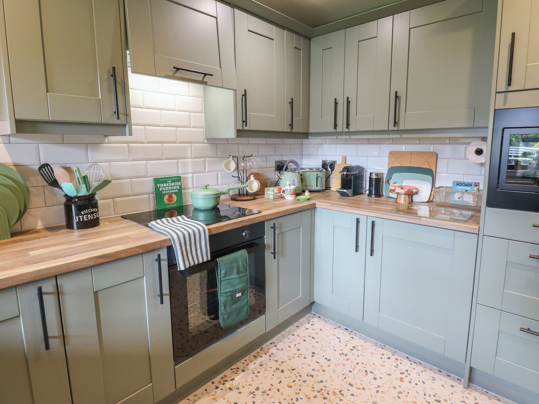 A kitchen with light green cabinets, a countertop with various appliances, and a backsplash.