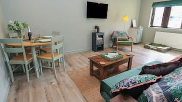 Ett vardagsrum med ett matbord i trä, gröna stolar, en soffa med mönstrade kuddar, en tv som är monterad på väggen och ett fönster med utsikt.