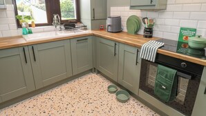 A kitchen with light green cabinets, a wooden countertop, a window with a view, and a green towel hanging on the oven door. - Underview (Holmfirth)