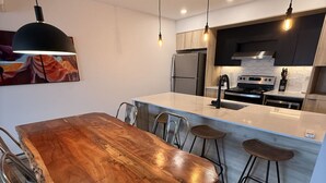 A modern kitchen with a wooden dining table, bar stools, stainless steel appliances, pendant lighting, and a tiled backsplash. - Modern 2BD/2BA Suite at Le Prisme w/ Indoor Pool, Gym & Yoga Studio (Québec)