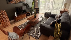 A modern living room with a sofa, coffee table, and TV stand. There is a large window with curtains, a plant, and a patterned rug. - Modern 2BD/2BA Suite at Le Prisme w/ Indoor Pool, Gym & Yoga Studio (Québec)